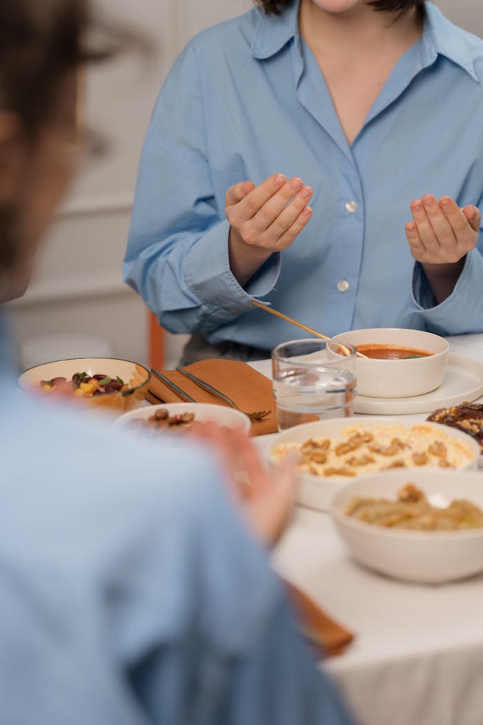Women in blue shirts are praying over a meal at a table, emphasizing faith and community.